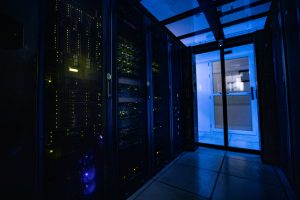 Server racks inside a dimly lit data center room with blue ambient lighting and glass doors leading to an adjacent control room